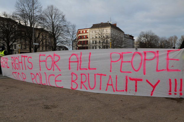 Gruppe von Menschen mit einem Banner "Rechte für alle Menschen Stoppt Polizeigewalt" vor einer Straßenlaterne, einem Schild, Bäumen, Gebäuden mit Fenstern und einem bewölkten Himmel im Hintergrund.