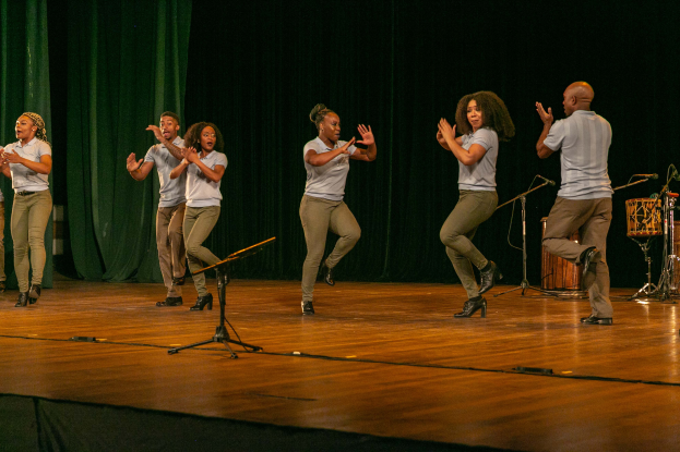 Eine Gruppe von Menschen auf einer Bühne mit Mikrofonen, Musikinstrumenten und einem Vorhang im Hintergrund, die Hände klappend feiern, die wie ein Tanzteam aussehen.