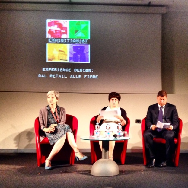Zwei Frauen und ein Mann in Anzügen sitzen auf roten Stühlen auf einer Bühne, mit einem Bildschirm dahinter und einem Tisch mit Wasserflaschen davor.