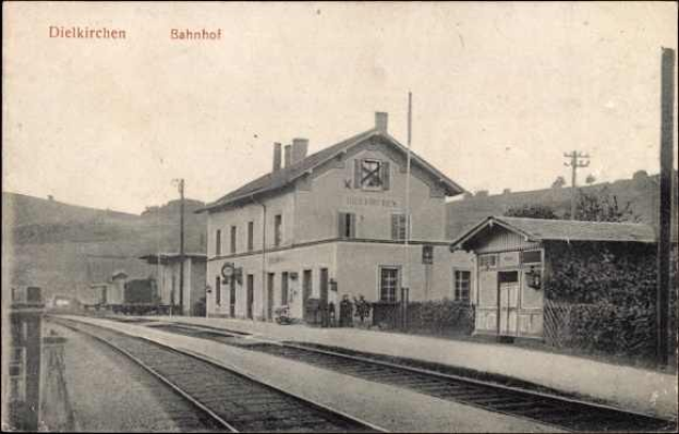 Schwarzes und weißes Foto einer Bahnstation in Dielkirchen, Deutschland, mit einem Zug auf den Gleisen, umgeben von Häusern, Strommasten, Bäumen, Menschen, Bergen im Hintergrund und einem klaren Himmel.