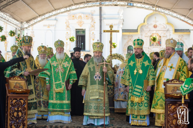 Gruppe von Priestern vor einer Kirche während einer religiösen Zeremonie, wobei einer ein Buch und ein Mikrofon hält, Kreuzsymbol und Blumen im Hintergrund.