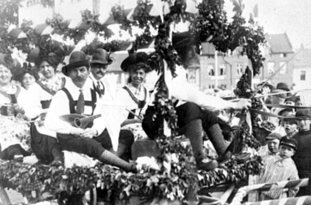 Eine Gruppe von Menschen in traditioneller bayrischer Tracht, einige mit Instrumenten, fährt auf einem mit Laub geschmückten Wagen in einem Umzug, mit Gebäuden und einem klaren Himmel im Hintergrund, dargestellt in Schwarz-Weiß.