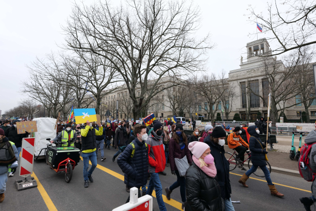 Eine große Gruppe von Menschen marschiert bei einer Demonstration auf einer Straße in Washington, D.C., mit Schildern und Bannern, einige fahren Fahrräder, unter einem klaren blauen Himmel vor einem Gebäude.