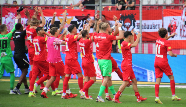 Group of soccer players celebrating a goal on a field, some holding bottles or clapping, with banners, a fence, chairs, and ceiling lights in the background.
