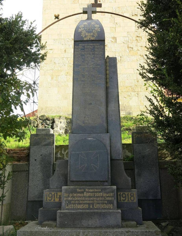 Ein steinernes Denkmal mit einem Kreuz, gewidmet den deutschen Soldaten, die im Zweiten Weltkrieg starben, steht vor einer Kirche mit einem kreuzförmigen Turm, umgeben von Bäumen und einer Mauer.