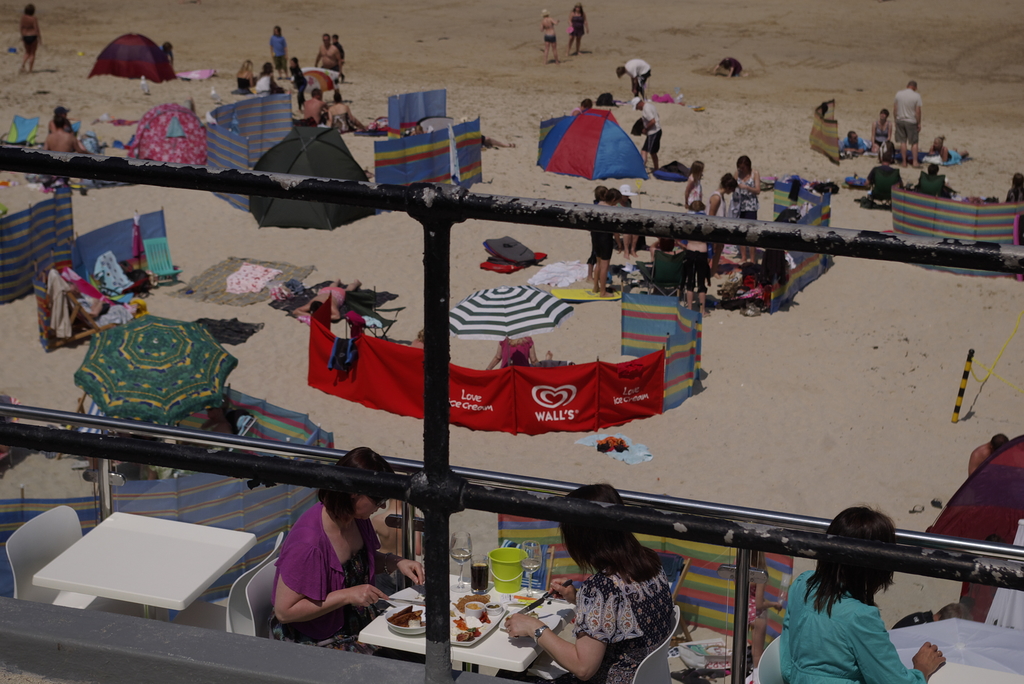 Eine schwarze Reling im Vordergrund mit einem leeren Tisch und Stühlen, zwei Frauen sitzen am Tisch und essen, Sand, ein rotes Tuch und einige Menschen stehen rechts im Hintergrund, mit Zelten und Kleidung weiter hinten.