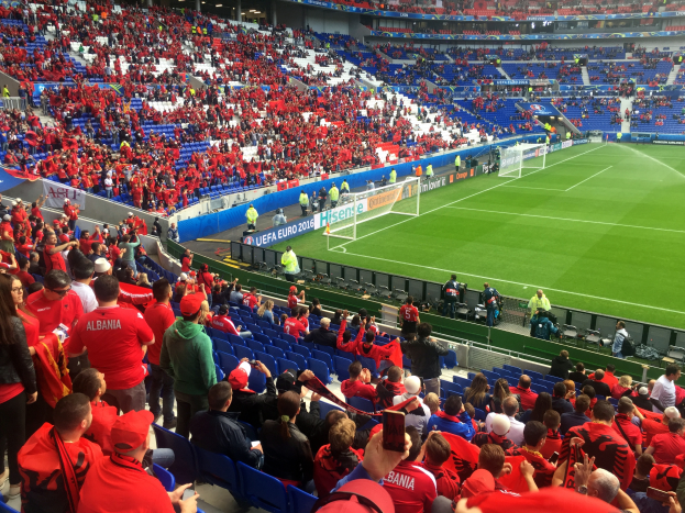 Ein vollbesetztes Stadion mit sitzenden und stehenden Zuschauern, Banner, Torpfosten und Deckenleuchten, während die Menge gespannt auf den Beginn eines Fußballspiels wartet.