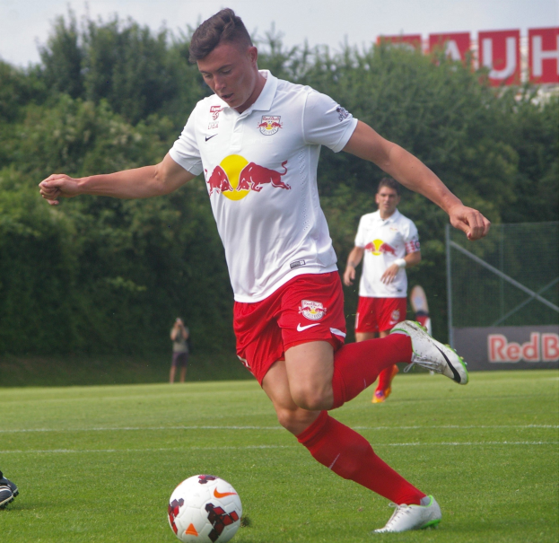 Ein professioneller Fußballspieler in einem roten Trikot kickt einen Ball auf einem grünen Feld mit Bäumen und einem klaren blauen Himmel im Hintergrund, in der Nähe eines Schildes mit der Aufschrift "RB Leipzig."