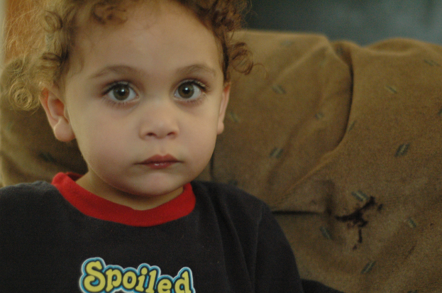 Ein Kind mit lockigen Haaren in einem schwarzen T-Shirt mit der gelben Aufschrift "Spoiled", das vor einem braunen Stoffhintergrund steht.