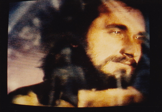 Ein Mann mit langem dunklen Bart und Haar, der ein weißes Hemd trägt, ist auf einem Fernseher zu sehen und schaut ernst und direkt.