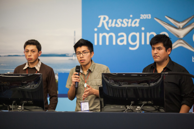 Drei Jungen stehen neben einem Schreibtisch mit zwei Computersystemen, einer spricht in ein Mikrofon und trägt eine ID-Karte mit roter Markierung, vor einer Wand mit einem Werbeplakat, auf dem 'Russia Imagine 2013' steht.