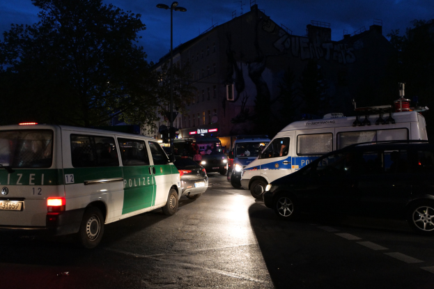 Eine Gruppe von Polizeiwagen, darunter ein Van, ein Auto und ein Rad, geparkt am Straßenrand bei Nacht, mit Gebäuden, Bäumen, Laternenpfählen und einem Sternenhimmel im Hintergrund.