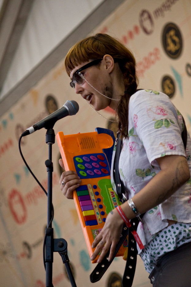 Eine Frau in einem weißen Hemd und Sonnenbrille singt in ein Mikrofon auf einem Ständer und hält eine Piano, mit einem Plakat an der Wand im Hintergrund.