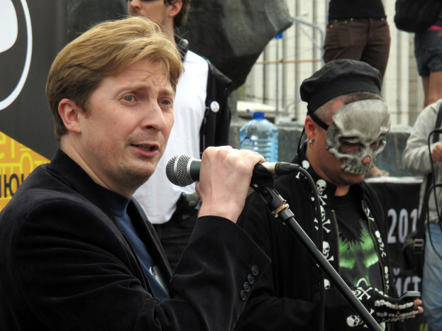 Ein Mann in einer schwarzen Jacke singt in ein Mikrofon auf der Bühne, hinter ihm stehen mehrere Personen, eine trägt eine Maske, und es gibt eine Wasserflasche und ein Banner im Hintergrund.