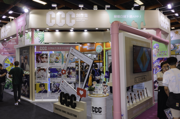 Eine Gruppe von Menschen vor einem Messestand mit Werbetafeln, Fernseher und Deckenbeleuchtung, wahrscheinlich eine kreative Comic-Sammlung.