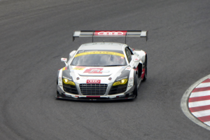 Ein leuchtend roter und weißer Audi R8 LMS GT3 Rennauto mit dem Audi-Logo auf der Seite, gesteuert von einer Person in einem schwarzen Rennanzug und Helm, fährt um eine Rennstrecke.