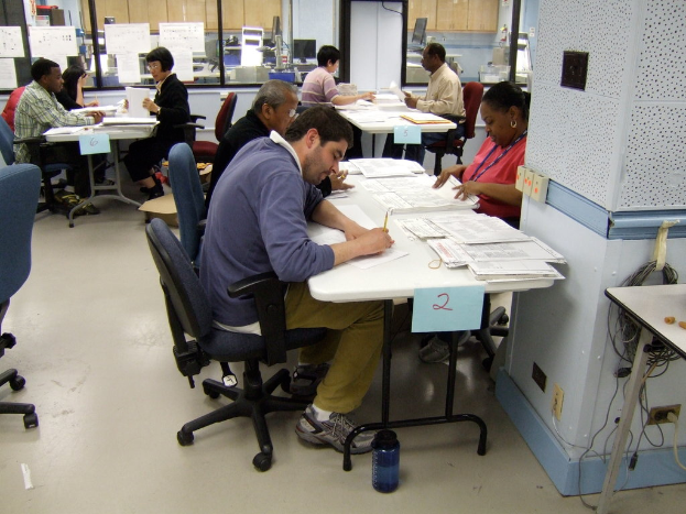 Eine Gruppe von Menschen, die an Schreibtischen sitzen und Papiere bearbeiten, einige schreiben und andere erklären, mit Glasfenstern und Monitoren im Hintergrund.
