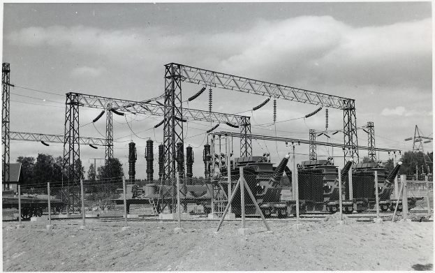 Ein Schwarz-Weiß-Foto einer elektrischen Umspannstation mit Transformatoren, Polen, Drähten, Maschinen, einem Zaun, Sand, Bäumen und einem bewölkten Himmel, mit Text unten.