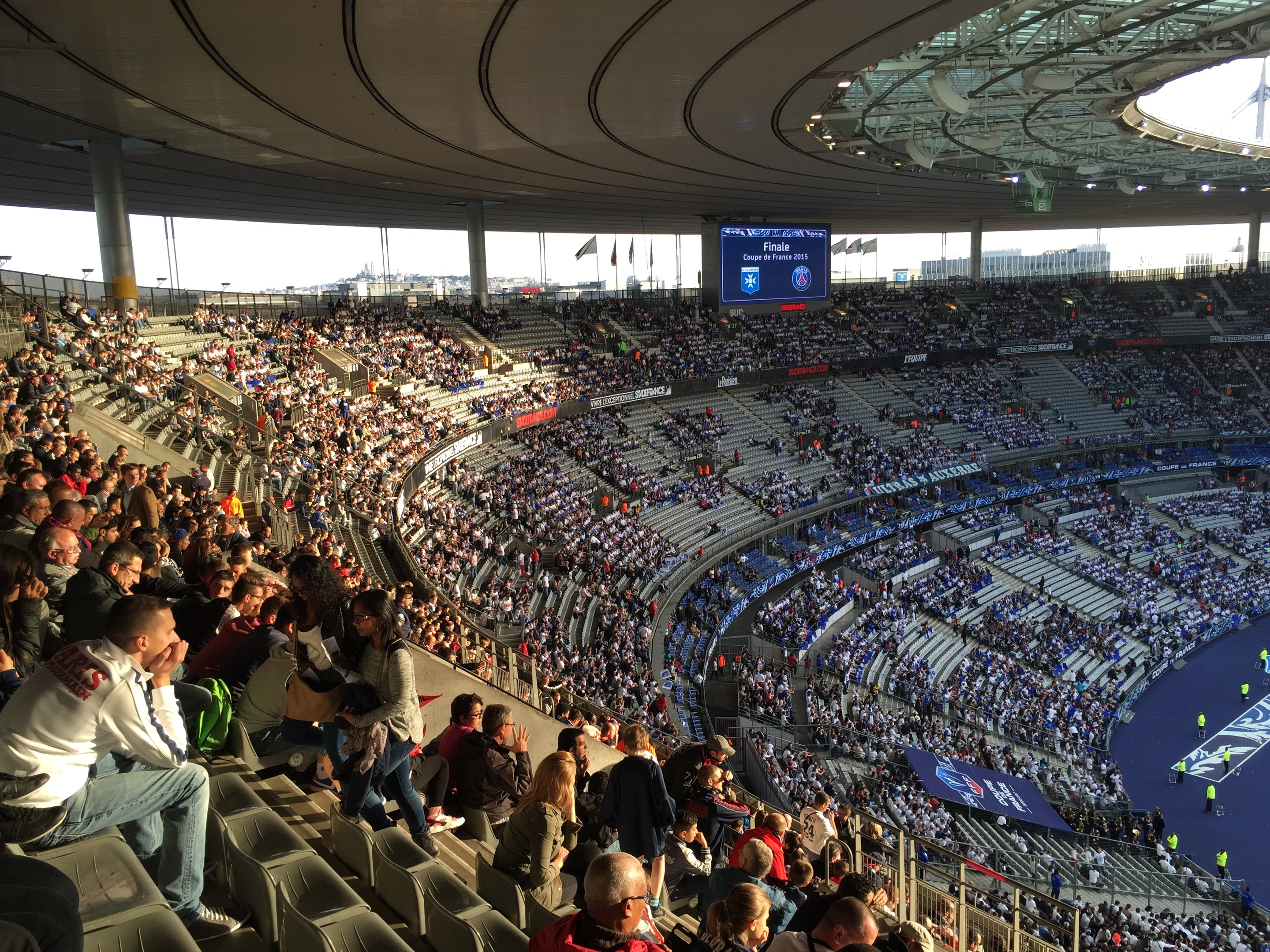 Große Menschenmenge in einem Stadion bei einem Fußballspiel mit einer Bühne, Fahnen und einem Hintergrundbild, identifiziert als Allianz Arena in München, Deutschland.