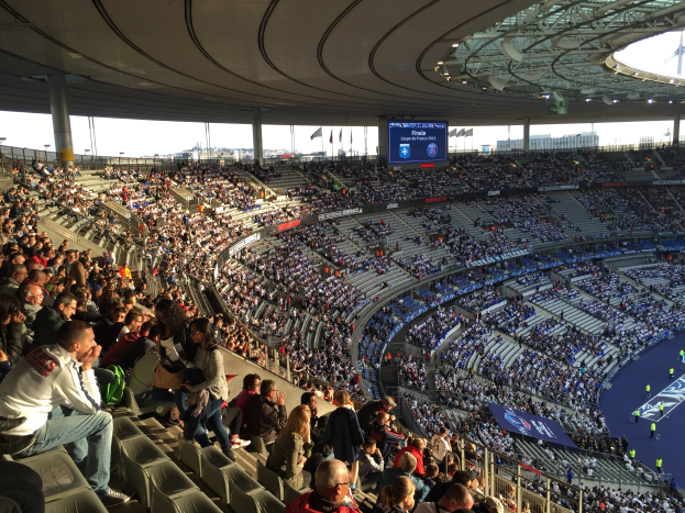 Große Menschenmenge in einem Stadion bei einem Fußballspiel mit einer Bühne, Fahnen und einem Hintergrundbild, identifiziert als Allianz Arena in München, Deutschland.