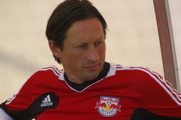 Ein Mann in einem roten und schwarzen T-Shirt sitzt auf einer Bank vor einer Wand und sieht selbstbewusst und entschlossen aus, wie ein professioneller Fussballspieler.