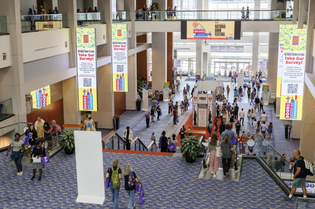 Eine große Menge bewegt sich durch ein großes Gebäude mit Bannern, Topfpflanzen, Rolltreppen, Geländern, Säulen, bedruckten Tafeln, Glasdoors und anderen Elementen, einige tragen Taschen, was auf den Besuch der Nationalen Buchmesse hinweist.