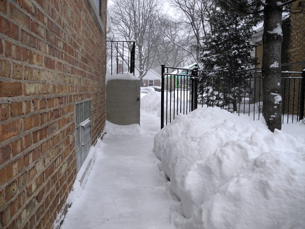 Eine verschneite Szene mit einer Wand auf der linken Seite, Schuppen, B├Ąume, Gitter und ein Netz im Hintergrund, alles mit Schnee ├╝berzogen, mit Schnee am Boden.