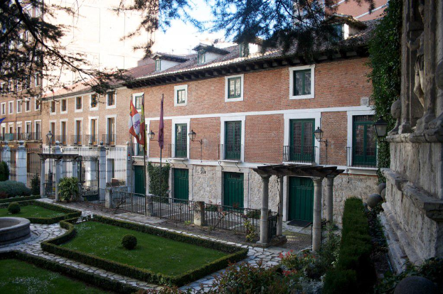 Mehrere Gebäude mit Flaggen, einem Zaun und Grünfläche davor, mit Pflanzen und einer Zaunwand in der rechten Ecke.