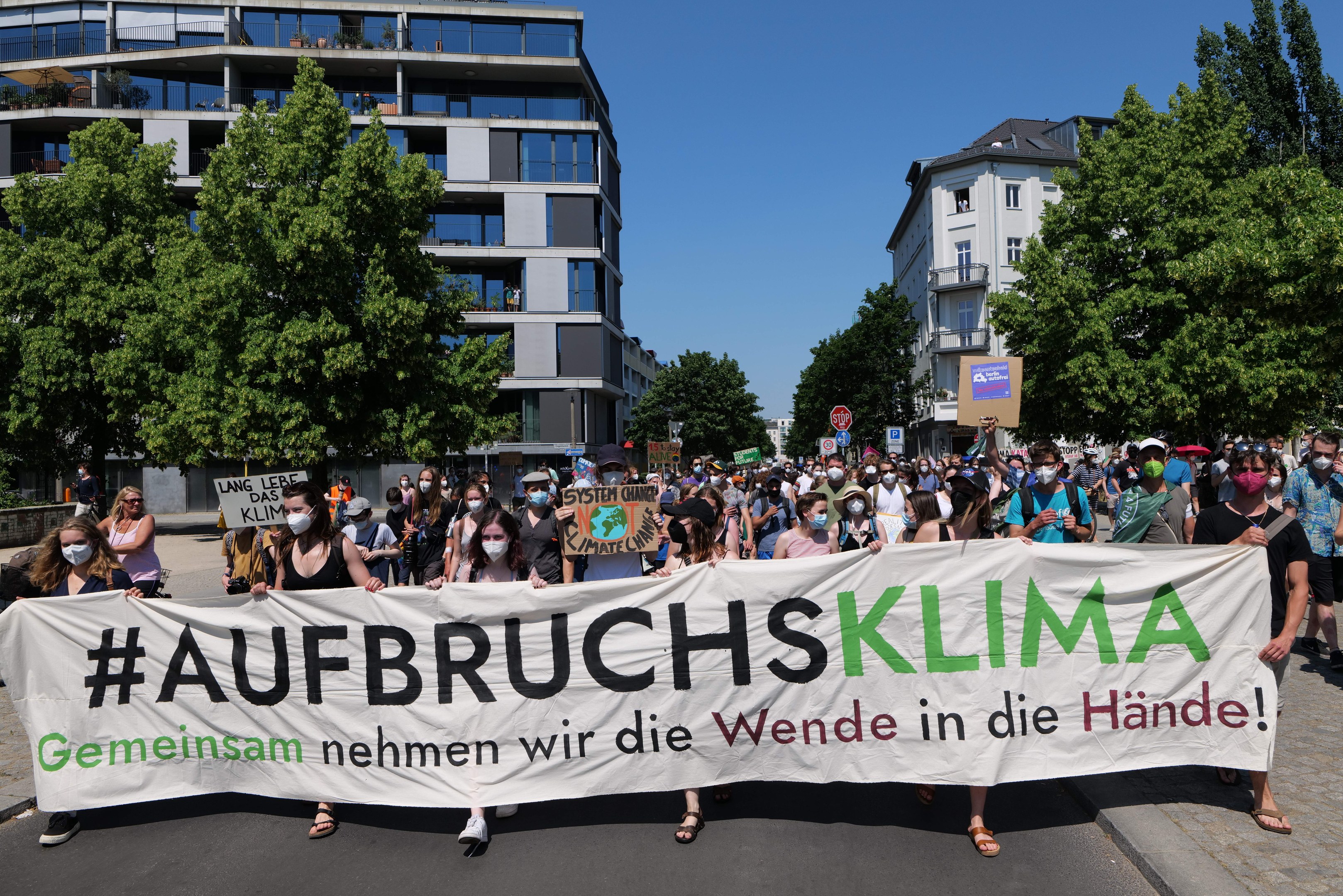 Gruppe von maskierten Menschen mit einem Banner 'Aufbruchsklima' vor einem Gebäude mit Bäumen und klarem Himmel.