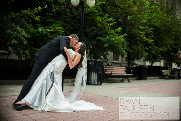 Eine Braut und ein Bräutigam küssen sich mit Bänken, Mülltonnen und Bäumen im Hintergrund.