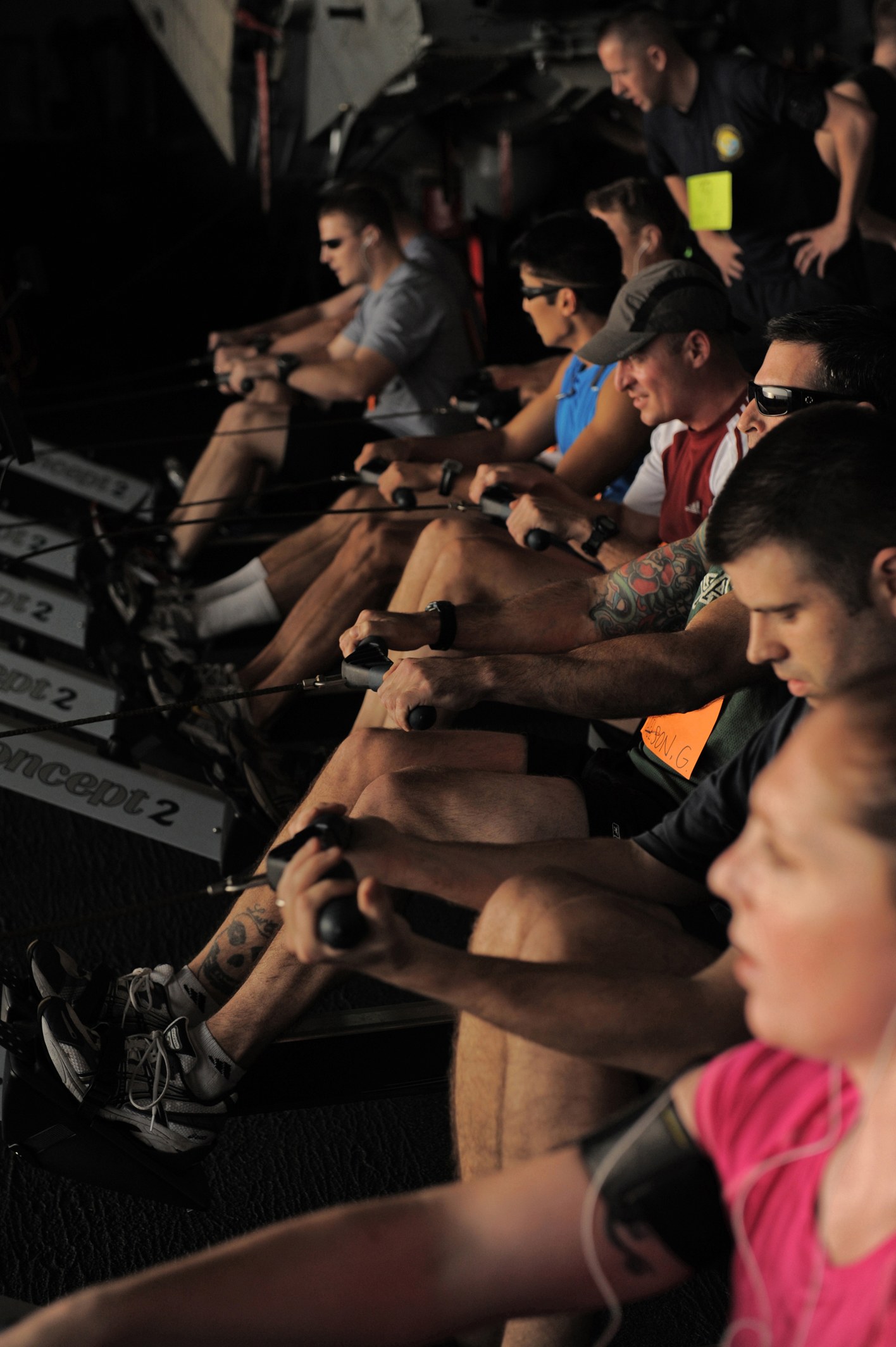 Eine Gruppe von Menschen, die Rudermaschinen in einem Fitnessstudio benutzen, mit anderen im Hintergrund, die alle sportliche Schuhe tragen.
