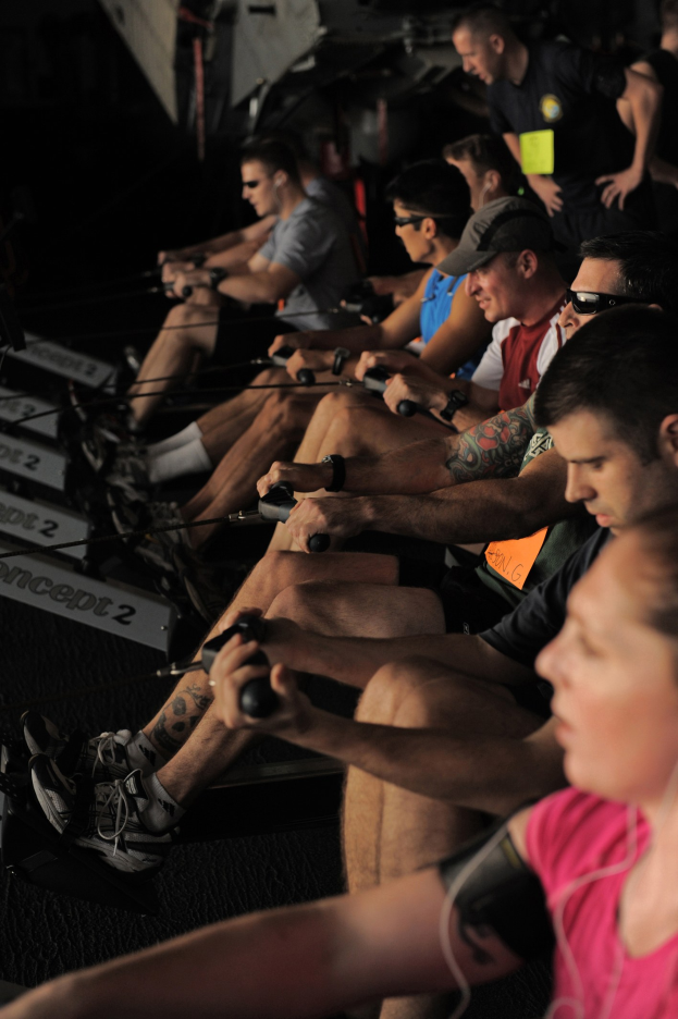 Eine Gruppe von Menschen, die Rudermaschinen in einem Fitnessstudio benutzen, mit anderen im Hintergrund, die alle sportliche Schuhe tragen.