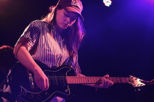 Eine Frau mit einer Mütze spielt Gitarre auf einer Bühne bei einem Musikkonzert, während zahlreiche Lichter den Hintergrund erhellen.