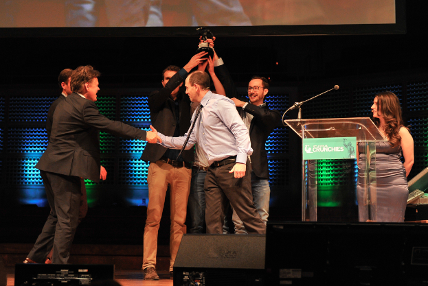 Zwei M├Ąnner in schwarzen Jacken heben einen Preis mit den H├Ąnden und l├Ącheln, w├Ąhrend ein Mann links die Hand sch├Ąktelt und eine Frau rechts steht.