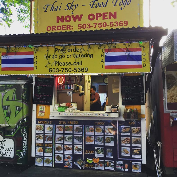 Ein Schaufenster mit verschiedenen Gegenständen wie Getränkedosen, Speisekarten, einer Uhr, Gewürzen in Plastikbehältern, Gewürzen, Werbetafeln, Namensschildern und einem Blick auf den Himmel.