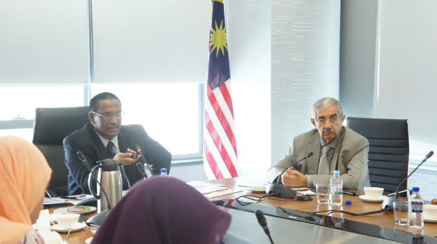 Eine Gruppe von Menschen, darunter Männer in Anzügen und zwei Frauen in Burkas, sitzt um einen Tisch mit Wasserflaschen, einem Mikrofon, Tassen und einem Teekessel; im Hintergrund ist ein Fenster mit Vorhängen und eine Flagge zu sehen.