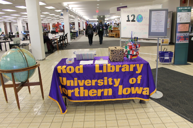 Eine Ausstellung mit einem Tisch, auf dem verschiedene Landesflaggen, eine Box, Papiere und ein Banner sowie links daneben eine Weltkugel, rechts daneben eine Anschlagtafel mit ein paar Menschen in der Nähe und links daneben mehrere Tische und Stühle zu sehen sind.