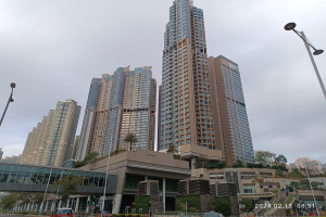 Eine Stadtstraße mit hohen Gebäuden, darunter ein markantes Wohnhaus im Vordergrund, Fahrzeuge, Laternenpfähle, eine Brücke, Bäume, Pflanzen und einen bewölkten Himmel.