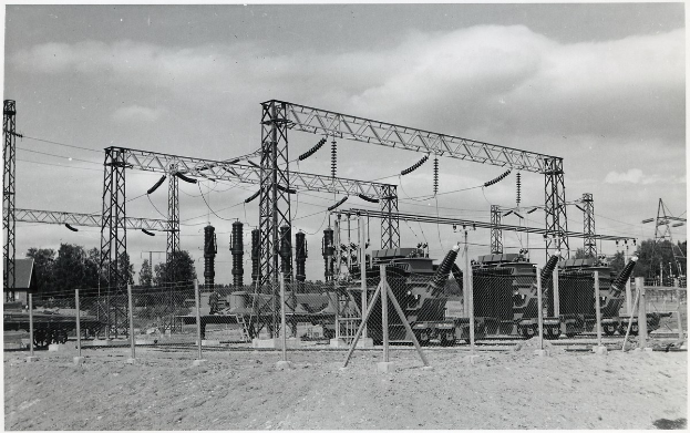 Schwarzes und weißes Foto einer elektrischen Umspannstation mit Transformatoren, Masten, Drähten, Maschinen, einem Zaun, Sand, Bäumen und einem bewölkten Himmel mit Text am unteren Rand.