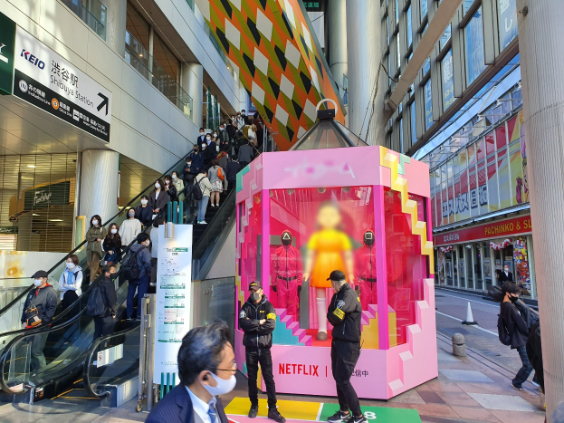 Eine Gruppe von Menschen steht vor einem Gebäude mit Glaswänden, Säulen und Texttafeln, einige tragen Masken, während eine Rolltreppe mit Menschen daneben führt; ein pinker Kasten in der Mitte zeigt Schaufensterpuppen und trägt den Text "Netflix's neuer Netflix-Stand in Tokyo."