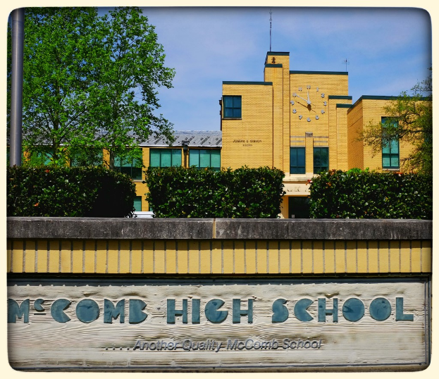 Ein Plakat mit der Aufschrift "MCCOMB HIGH SCHOOL" an einer Wand, umgeben von Pflanzen, Bäumen, einem Pfahl, einem Gebäude und einem bewölkten Himmel.
