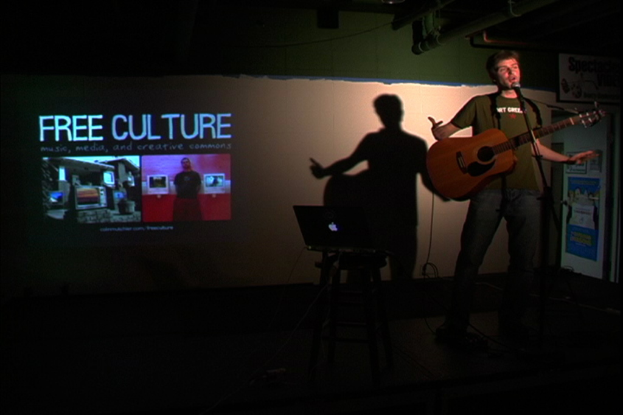 Eine Person, die eine Gitarre hält und auf einem Tisch ein Laptop und im Hintergrund ein Projektionsbild mit der Aufschrift "Free Culture" zu sehen ist.