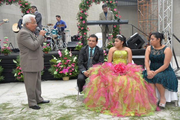 Ein Mann steht und spricht in ein Mikrofon, während andere sitzen und ein Orchester im Hintergrund auf einer Bühne spielt, die mit Blumen und Pflanzen geschmückt ist.