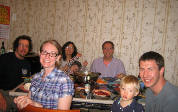 Ein Esstisch voller Gerichte und Essen in einem Wohnzimmer mit Blumenmuster an den Wänden, umgeben von fünf lächelnden Menschen, mit einem Kalender und einer Flasche an der Wand und einem Stuhl im Vordergrund.