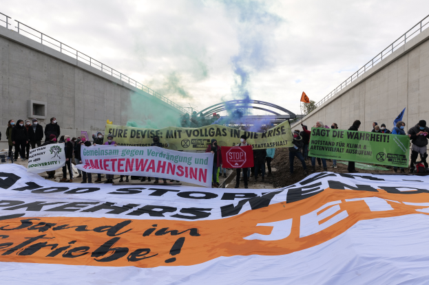 Eine Gruppe von Menschen steht vor einem großen Banner mit der Aufschrift "Klima-Protest in Berlin", hält Schilder und Fahnen hoch, mit Rauch im Hintergrund und einem bewölkten Himmel darüber.