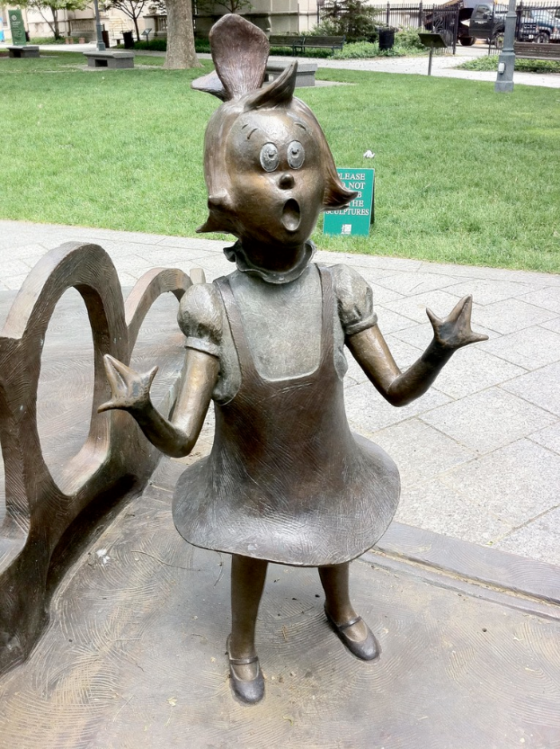 Eine Metallskulptur eines Mädchens steht auf einer grünen Fläche mit Bäumen, Mülltonnen, einem Metallzaun, parkenden Autos und Gebäuden im Hintergrund.