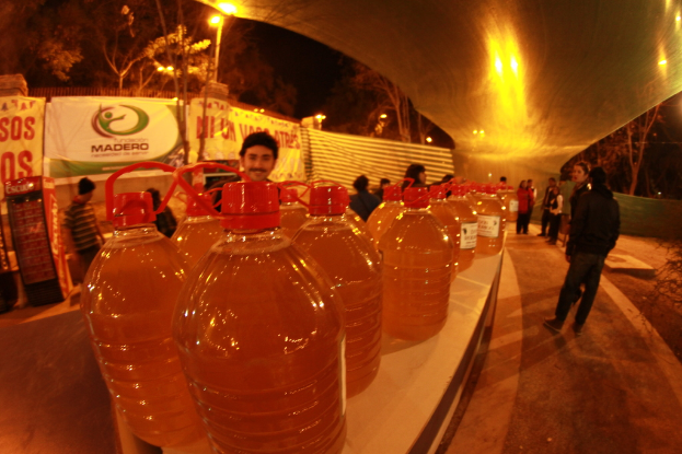 Flaschen mit roten Kappen, die mit Flüssigkeit gefüllt sind, werden auf einem Tisch platziert, hinter denen Menschen stehen und Schilder im Hintergrund zu sehen sind, von denen eines 'mandero' lautet, und weiter hinten Bäume.