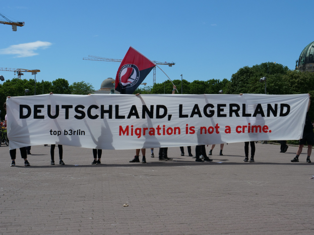 Gruppe von Menschen mit einem Banner "Deutschland, Lagerland Migration ist kein Verbrechen" und einer Flagge, mit Straßeninfrastruktur und Gebäuden im Hintergrund.