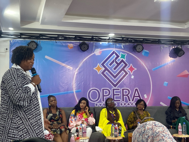 Frau steht vor sitzenden Frauen auf einer Couch, hält ein Mikrofon, mit Flaschen und Gegenständen auf den Tischen im Vordergrund, ein Banner mit Text und Deckenbeleuchtung im Hintergrund.