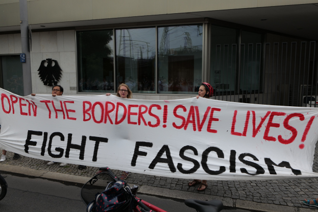 Gruppe von Menschen mit einem Transparent "Öffnet die Grenzen, rettet Leben, kämpft gegen Faschismus" vor einem Gebäude mit Glasfenstern und einer Stange, mit einem Fahrrad und einer Tasche auf der Straße.
